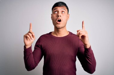 Beyaz izole edilmiş arka plan üzerinde duran genç ve yakışıklı İspanyol adam şaşırmış ve şaşırmış bir şekilde yukarı bakıp parmaklarıyla işaret ediyor ve kollarını kaldırıyordu..