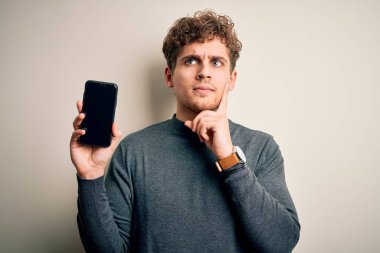Sarışın, kıvırcık saçlı, akıllı telefon gösteren, soyutlanmış beyaz arka planda duran, soru soran, kafası karışık bir genç adam.