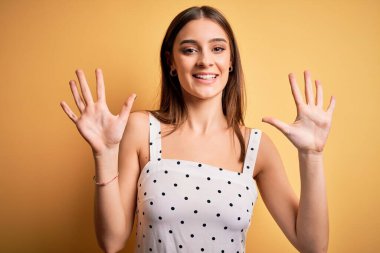 Sarı arka planda dikilen, rahat elbiseler giyen genç, güzel esmer kadın on numarayı gösterirken kendinden emin ve mutlu bir şekilde gülümsüyor..