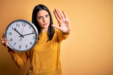 Mavi gözlü genç bir kadın geri sayım yapıyor sarı arka planda büyük bir saat tutuyor açık elle dur işareti yapıyor ciddi ve kendinden emin bir ifadeyle savunma hareketi yapıyor.