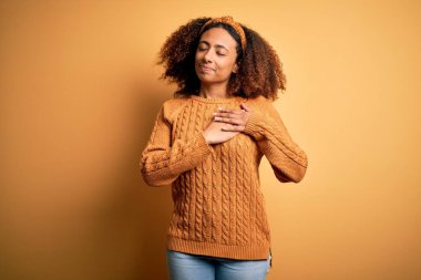Afro saçlı, sarı arka planda rahat süveter giyen, elleri göğsünde, gözleri kapalı ve yüzünde minnet dolu bir jest olan Afro kökenli genç bir Amerikalı kadın. Sağlık kavramı.