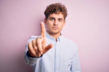 Sarışın, kıvırcık saçlı, beyaz arka planda çizgili tişört giyen, parmağını kaldırmış ve kızgın bir ifade sergileyen genç bir adam.