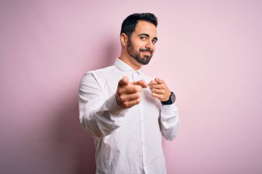 Genç, sakallı, rahat gömlekli pembe arka planda durup mutlu ve komik yüzlü parmakları kameraya gösteren yakışıklı bir adam. İyi enerji ve titreşimler.