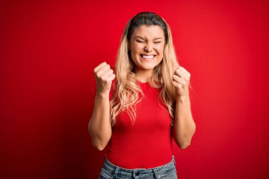 İzole edilmiş kırmızı arka planda dikilen ve kolları havada, gözleri kapalı zaferi kutlayan gündelik tişört giyen genç, güzel sarışın kadın. Kazanan konsept.