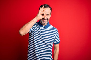 Orta yaşlı, beyaz, sıradan çizgili polo giyen, izole edilmiş kırmızı arka planda duran, el hareketleriyle gülümseyen, mutlu yüzüyle parmaklarının arasından bakan bir adam..