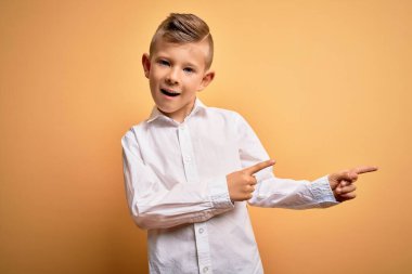 Mavi gözlü, sarı arka planda zarif beyaz gömlek giyen, gülümseyen ve kameraya bakıp iki eliyle ve parmaklarıyla yüzünü gösteren küçük beyaz bir çocuk..