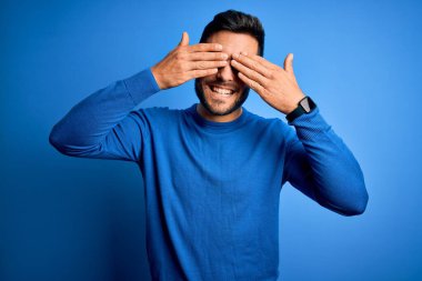 Sakallı, sıradan süveter giyen, mavi arka planda gözlük takan, elleri neşeli ve komik gülümseyen genç yakışıklı bir adam. Kör kavram.
