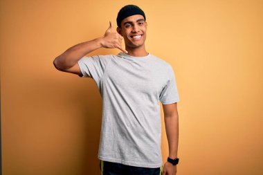 Yakışıklı Afro-Amerikan sporcusu sarı arka planda spor kıyafetleri giyip telefonla el ve parmak hareketleriyle telefonla konuşuyor gibi gülümsüyor. İletişim kavramları.
