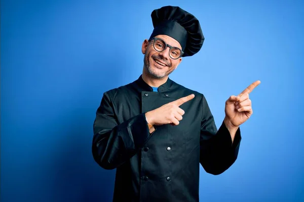 若いですハンサムなシェフ男身に着けています炊飯器制服と帽子上の隔離された青の背景笑顔と見ますカメラポインティングで二本の手と指で側面 — ストック写真