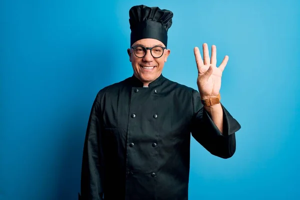 Middle age handsome grey-haired chef man wearing cooker uniform and hat showing and pointing up with fingers number four while smiling confident and happy.