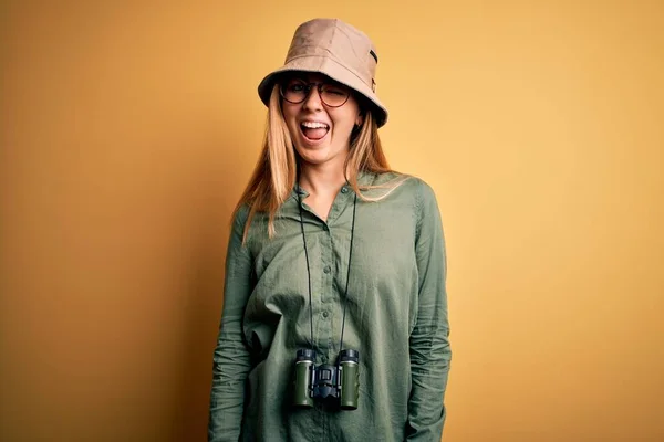 Beautiful blonde explorer woman with blue eyes wearing hat and glasses using binoculars winking looking at the camera with sexy expression, cheerful and happy face.