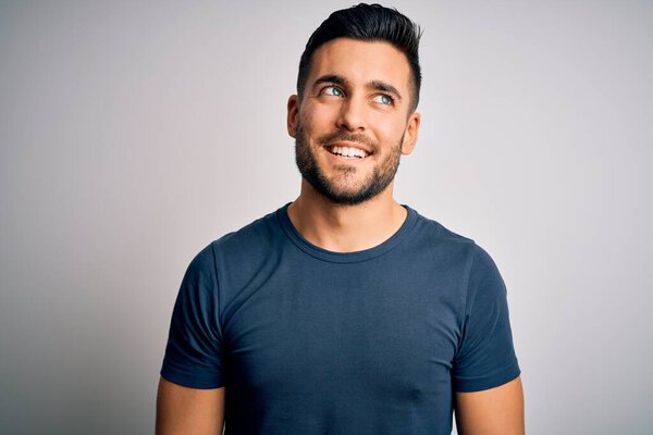 Young handsome man wearing casual t-shirt standing over isolated white background looking away to side with smile on face, natural expression. Laughing confident.