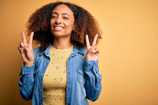 黄色の背景の上にカジュアルなデニムシャツを着てアフロの髪を持つ若いアフリカ系アメリカ人の女性は 勝利のサインを行う指を示すカメラを見て微笑んだ 第二番 — ストック写真