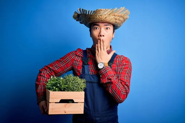 Young handsome chinese farmer man wearing apron and straw hat holding box with plants cover mouth with hand shocked with shame for mistake, expression of fear, scared in silence, secret concept