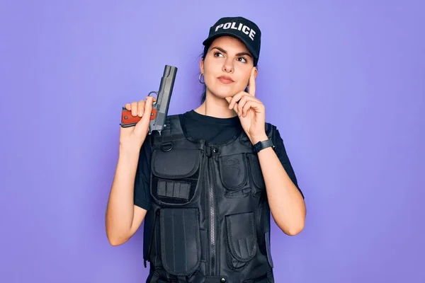 Young police woman wearing security bulletproof vest uniform and holding gun serious face thinking about question, very confused idea