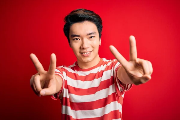 双方の手の指が勝利記号を行うことを示す舌で笑顔の赤い背景の上に立ってカジュアルな縞模様のTシャツを着て若いハンサムな中国人男性 第二番 — ストック写真