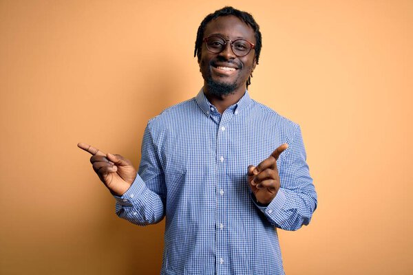 Young handsome african american man wearing shirt and glasses over yellow background smiling confident pointing with fingers to different directions. Copy space for advertisement
