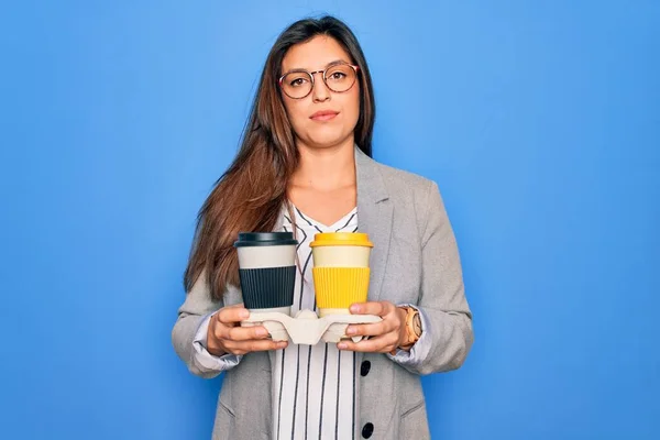 Young hispanic intern business woman holding coffee over blue background with a confident expression on smart face thinking serious