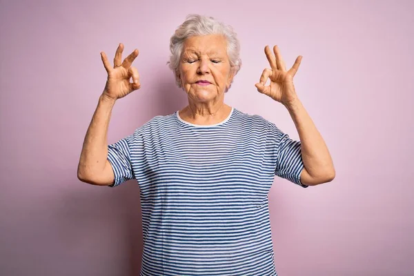 Senior beautiful grey-haired woman wearing casual t-shirt over isolated pink background relax and smiling with eyes closed doing meditation gesture with fingers. Yoga concept.