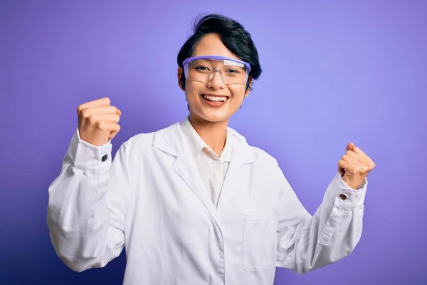 紫色の背景にコートと眼鏡をかけた若い美しいアジアの科学者の女の子は 腕を上げて目を開けて成功したことに驚きと驚きを祝います 勝者のコンセプト — ストック写真