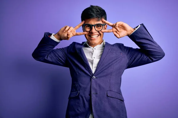 Young handsome business man wearing jacket and glasses over isolated purple background Doing peace symbol with fingers over face, smiling cheerful showing victory