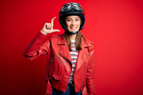 若い美しいブルネットのオートバイのヘルメットと赤いジャケットを身に着けているオートバイの女性は笑顔と指を探しているとカメラと小さなサイズの看板を行う手で自信を持ってジェスチャー 概念の測定 — ストック写真