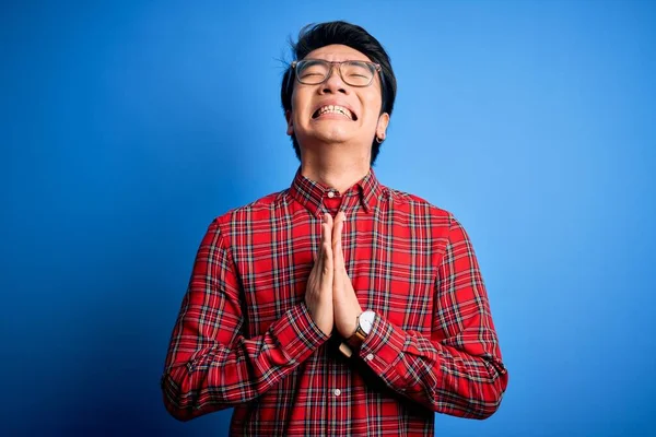 Young handsome chinese man wearing casual shirt and glasses over blue background begging and praying with hands together with hope expression on face very emotional and worried. Begging.