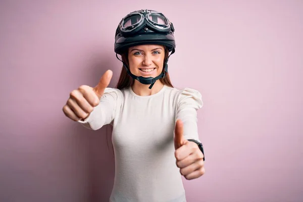 手で積極的なジェスチャーを行うことを承認ピンクの背景に元ヘルメットを身に着けて青い目を持つ若い美しいオートバイの女性 親指アップ笑顔と成功のために幸せ 勝者のジェスチャー — ストック写真
