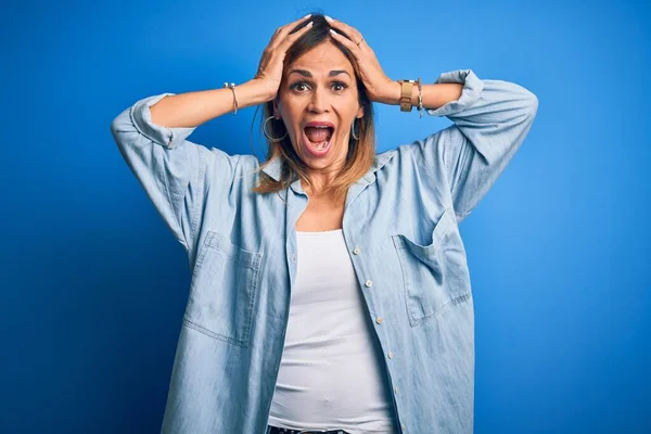 Middle age beautiful woman wearing casual shirt standing over isolated blue background Crazy and scared with hands on head, afraid and surprised of shock with open mouth