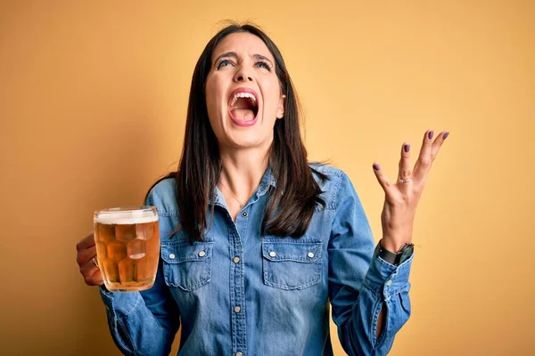 青い目の若い女性は ビールの瓶を飲んでいる独立した黄色の背景の上に立って狂気と怒りの叫びと積極的な表現と腕を上げて叫んでいた 欲求不満の概念 — ストック写真