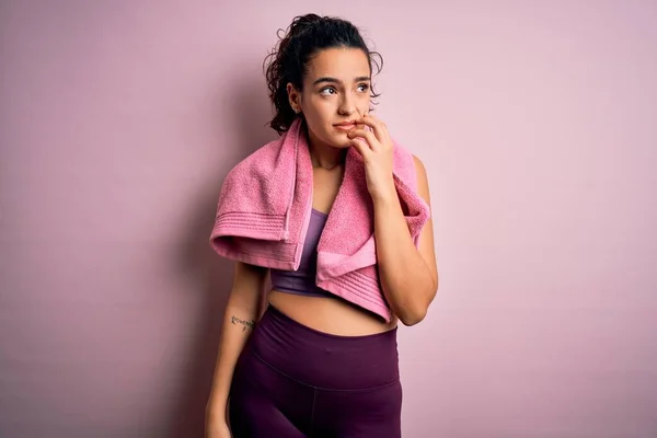Young beautiful sportswoman with curly hair doing sport using towel over pink background looking stressed and nervous with hands on mouth biting nails. Anxiety problem.