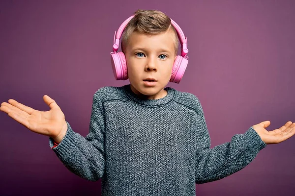 幼い白人の子供は 紫色の背景に音楽を聞いてヘッドフォンを着用し 腕や手を上げて混乱した表現をしていました 疑いの概念 — ストック写真