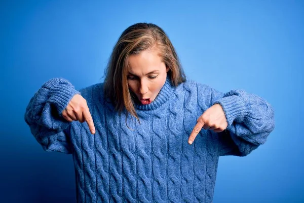 Young beautiful blonde woman wearing casual turtleneck sweater over blue background Pointing down with fingers showing advertisement, surprised face and open mouth