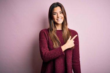 İzole edilmiş pembe arka planda rahat bir süveter giyen genç ve güzel bir kız. Yüzünde mutlu ve doğal bir ifadeyle el ve parmak uçlarını gösteren bir gülümseme var.