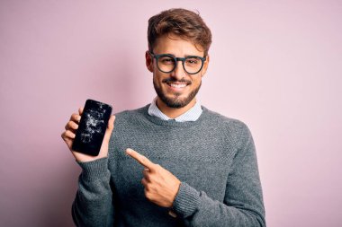 Sakallı, gözlüklü, kırık ve çatırdayan akıllı telefonlu genç adam elini ve parmağını göstererek çok mutlu oluyor.