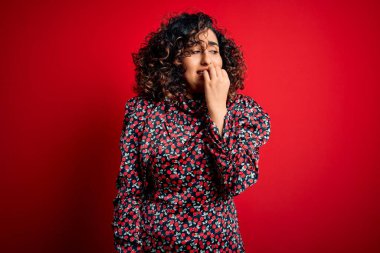 Genç, kıvırcık Arap bir kadın kırmızı arka planda dikilen sıradan bir çiçek elbisesi giyiyor. Elleri ağızdan tırnak yerken gergin ve stresli görünüyor. Endişe sorunu.