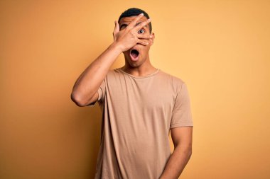 Genç, yakışıklı, Afro-Amerikan bir adam günlük tişört giyiyor sarı arka planda dikiliyor, yüzünü ve gözlerini eliyle kapatıyor, utanmış bir ifadeyle parmaklarından bakıyor..