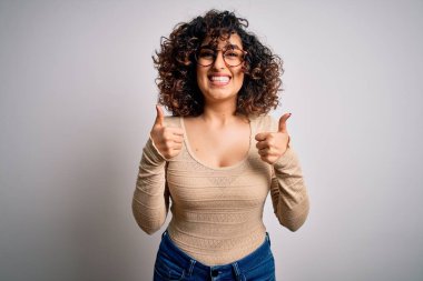 Güzel kıvırcık Arap kadın beyaz arka plan işareti üzerine günlük tişört ve gözlük takıyor. Elleriyle olumlu jest yapıyor. Gülümsüyor ve mutlu. Neşeli ifade ve kazanan jest.