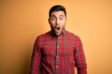 Sarı arka plan üzerinde duran rahat tişörtlü genç yakışıklı adam korku, korku ve heyecan dolu yüz ifadesiyle korkmuş ve şok olmuştu..