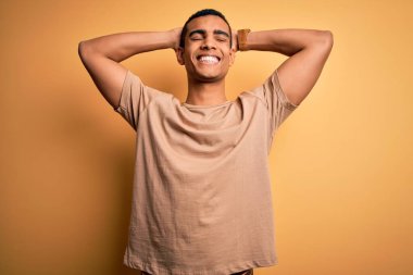 Genç, yakışıklı, Afro-Amerikalı bir adam, günlük tişört giyiyor sarı arka planda duruyor, rahatlıyor ve esniyor, kolları ve elleri başının arkasında, gülümsüyor.