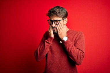 Sakallı, gözlüklü ve süveterli genç bir adam kırmızı arka planda duruyor. Yorgunluk ve baş ağrısı için gözlerini ovuşturuyor, uykulu ve yorgun bir ifade takınıyor. Görüş sorunu