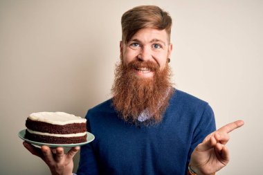 Doğum günü pastası tutan sakallı kızıl saçlı İrlandalı bir adam soyutlanmış arka planda süslemeleri olan çok mutlu bir şekilde elini ve parmağını kenarı işaret ediyor.