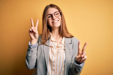 İzole edilmiş sarı arka planda gözlüklü, ceketli, güzel kızıl saçlı kadın kameraya bakıp zafer işareti yaparken gülümsüyor. İki numara..