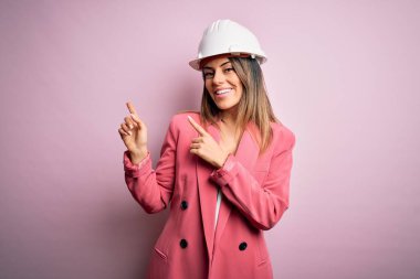 Pembe arka planda güvenlik kaskı takan genç ve güzel mimar kadın gülümsüyor ve kameraya bakıyor iki eliyle ve parmaklarıyla kenarı işaret ediyor..