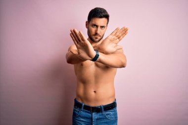 Sakalsız, genç, yakışıklı, güçlü bir adam pembe arka planda duruyor. Kollarını ve avuçlarını çapraz yapıp negatif işaret veriyor. Kızgın surat.