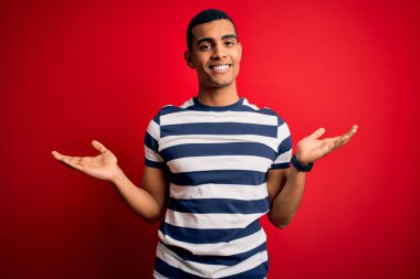 Yakışıklı Afro-Amerikan erkeği kırmızı arka planda dikilen günlük çizgili tişört giyiyor. Gülümsüyor, iki elini de açık gösteriyor. Karşılaştırma ve reklam karşılaştırması yapıyor ve dengeyi sağlıyor.