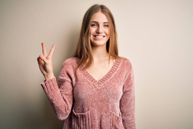 İzole edilmiş beyaz arka planda pembe kazak giyen güzel, kızıl saçlı kadın kameraya bakıp zafer işareti yaparken gülümsüyor. İki numara..