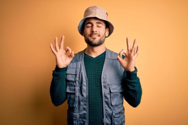 Tatilde sakallı yakışıklı turist sarı arka planda kaşif şapkası takıyor rahatlamış ve gözleri kapalı meditasyon hareketi yaparken parmaklarıyla gülümsüyor. Yoga konsepti.