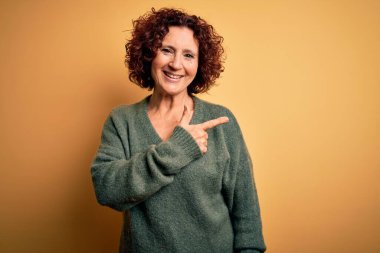 Orta yaşlı güzel kıvırcık saçlı kadın, izole edilmiş sarı arka plan üzerine gündelik süveter giyer. Neşeli, yüzünde mutlu ve doğal bir ifadeyle, elleri ve parmaklarıyla yüzünü gösteren bir gülümseme.