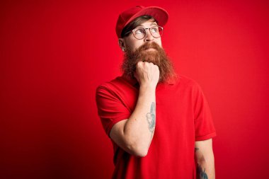 Gözlüklü yakışıklı teslimatçı ve izole edilmiş arka planda kırmızı şapkalı, elleri çenesinde soru sorarken, düşünceli bir yüz ifadesi vardı. Gülen ve düşünceli bir yüz. Şüphe kavramı.